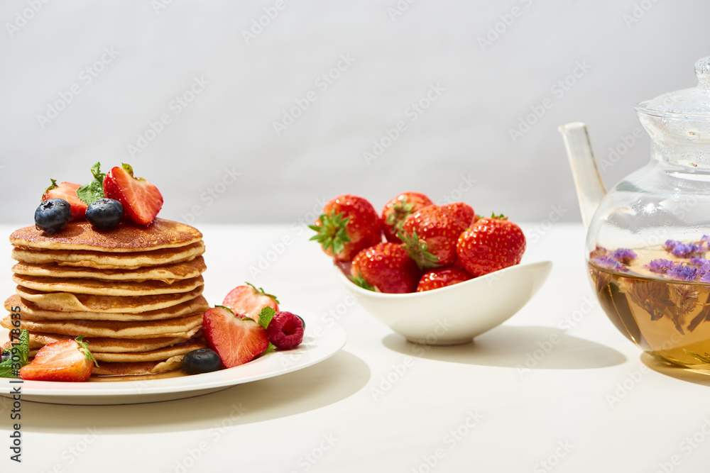 delicious pancakes with blueberries and strawberries on plate near herbal tea in teapot on white surface isolated on grey delicious pancakes with blueberries and strawberries on plate near herbal tea in teapot on white surface isolated on grey