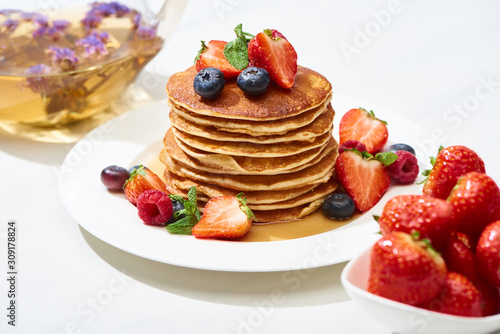 selective focus of delicious pancakes with honey, blueberries and strawberries on plate on white surface