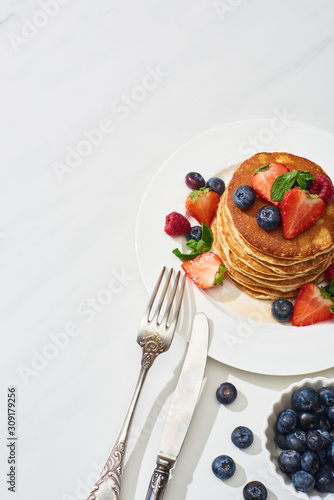 top view of delicious pancakes with maple syrup  blueberries and strawberries on plate near fork and knife on marble white surface