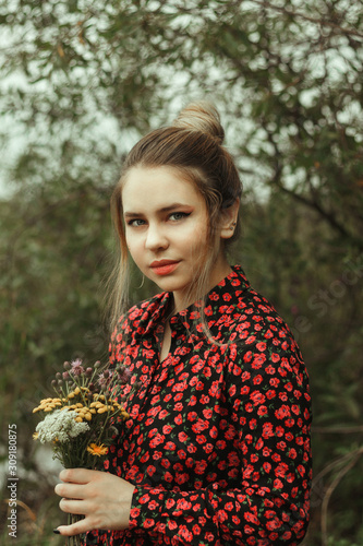 A young woman in a black-red dress with her hair gathered in a bun. In her hands holds a bouquet of wildflowers. A girl stands in a green meadow by the river.