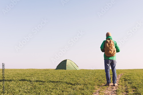 Tourist with backpack going to his tent on summer field. Travel concept