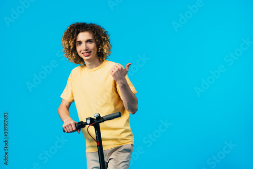 smiling curly teenager riding electric scooter and showing thumb up isolated on blue © LIGHTFIELD STUDIOS