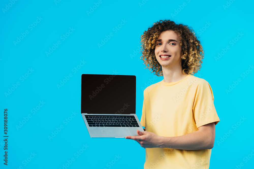 smiling curly student holding laptop with blank screen isolated on blue smiling curly student holding laptop with blank screen isolated on blue