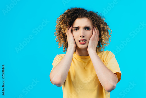 stressed curly teenager covering ears with hands isolated on blue