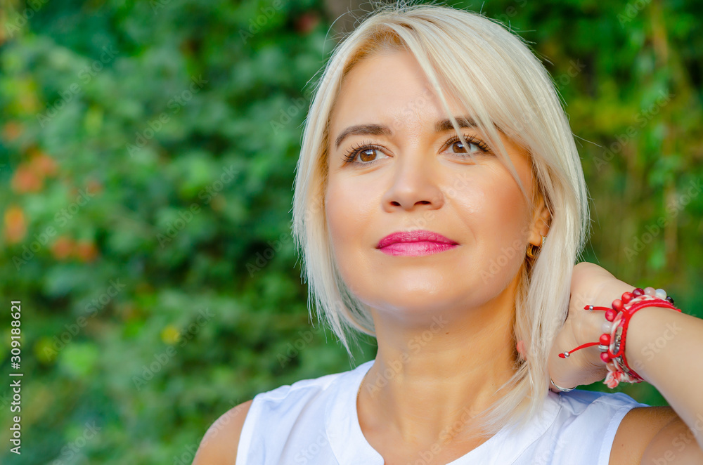 Beautiful blonde middle-aged woman in green leaves background. In white blouse, standing on street. Pretty smile on face straight short hair, brown eyes Beautiful blonde middle-aged woman in green leaves background. In white blouse, standing on street. Pretty smile on face straight short hair, brown eyes