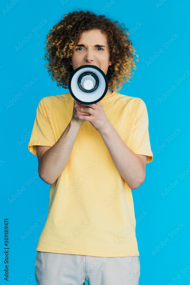 Obraz premium angry curly teenager screaming in loudspeaker isolated on blue
