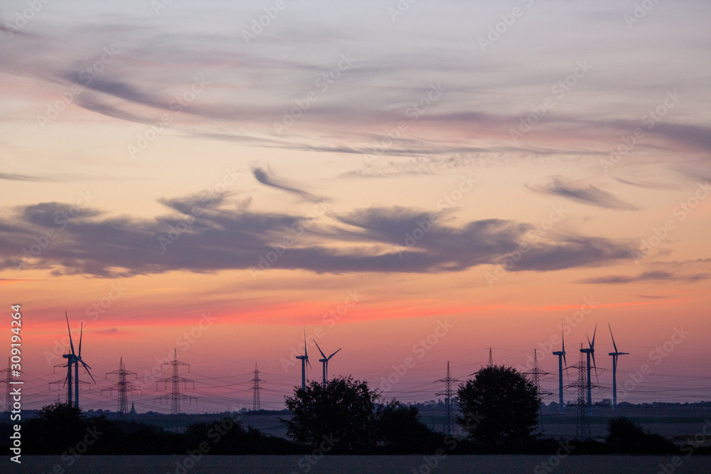 Wind farm with electricity pylons at dusk and red cloud sky