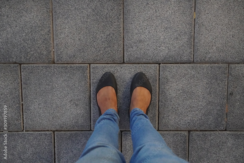 Women Black casual shoes standing and resting on asphalt concrete floor with square tiles. Top View. Concrete floor texture pattern pavement background. Selfie Female of Feet and Legs Seen from Above. Women Black casual shoes standing and resting on asphalt concrete floor with square tiles. Top View. Concrete floor texture pattern pavement background. Selfie Female of Feet and Legs Seen from Above.