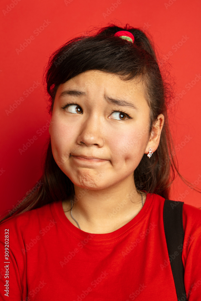 Asian teenager's portrait isolated on red studio background. Beautiful female brunette model with long hair in casual style. Concept of human emotions, facial expression, sales, ad. Uncertainty. Asian teenager's portrait isolated on red studio background. Beautiful female brunette model with long hair in casual style. Concept of human emotions, facial expression, sales, ad. Uncertainty.