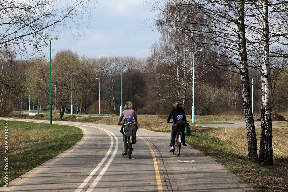 Two women with children go by bicycles on a bicycle track in the spring park Two women with children go by bicycles on a bicycle track in the spring park