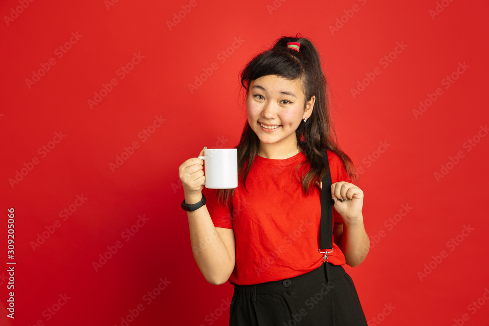 Naklejka premium Asian teenager's portrait isolated on red studio background. Beautiful female brunette model with long hair in casual style. Concept of human emotions, facial expression, sales, ad. Drinks tea, coffee