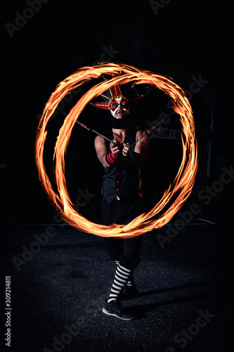Man in red jester costume with double fire chains performing swing make flame in circle moment overlap in center in the dark