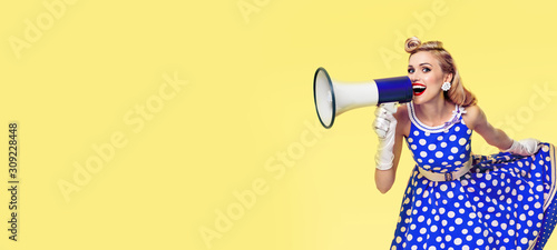 Retro, vintage and holiday sales concept picture- Woman holding megaphone, dressed in pin up style blue dress, over yellow background. Caucasian blond model - retro fashion.