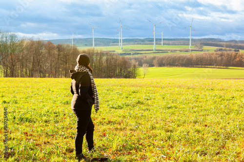 Lady is looking at the landscape with some wind turbine towers while standing in an open field during a cold winter afternoon.