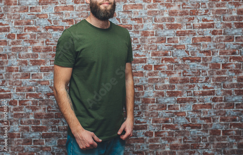 City portrait of handsome hipster guy with beard wearing a blank green military t-shirt standing on a brick wall background. Empty space for your logo or design. Mockup for print.