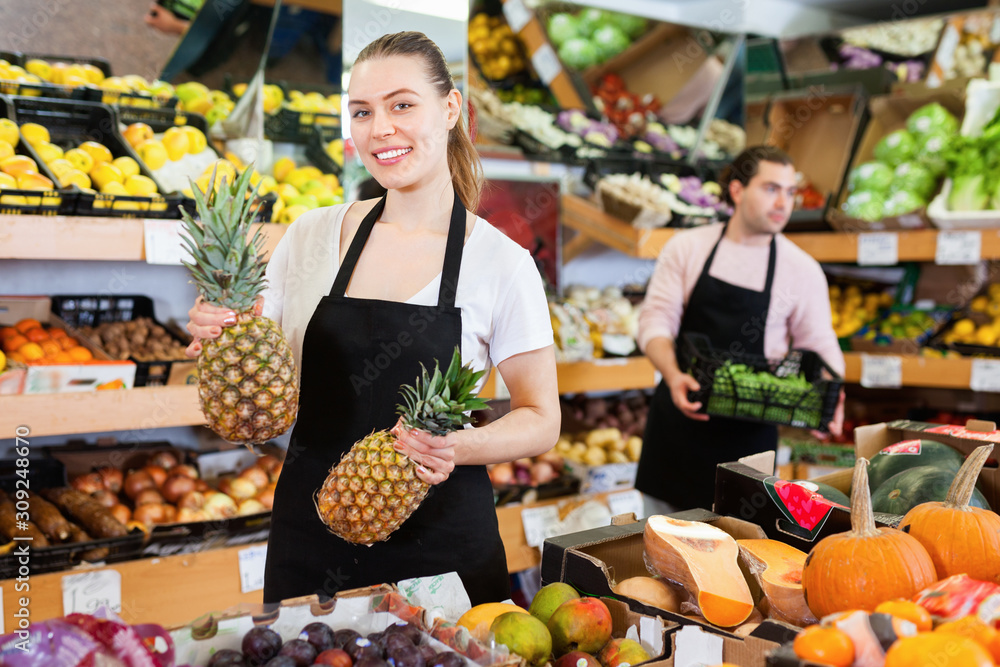 Positive saleswoman in apron offering fresh pineapples on the fruit store