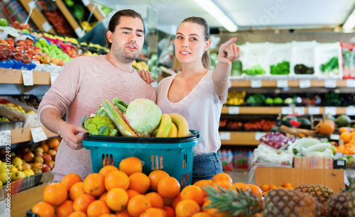 Positive family couple standing with full cart after shopping and pointing to shelves in fruit store