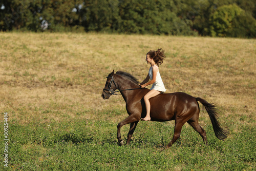 Young girl in white dress running on horse without saddle through meadow on summer afternoon