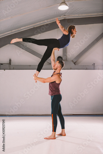 The theme of Acroyoga and Yoga Poses. A pair of two men and a woman stand in the position of asana. The guy holds the girl arched high back on the outstretched arm. In the gym with a studio Backlight
