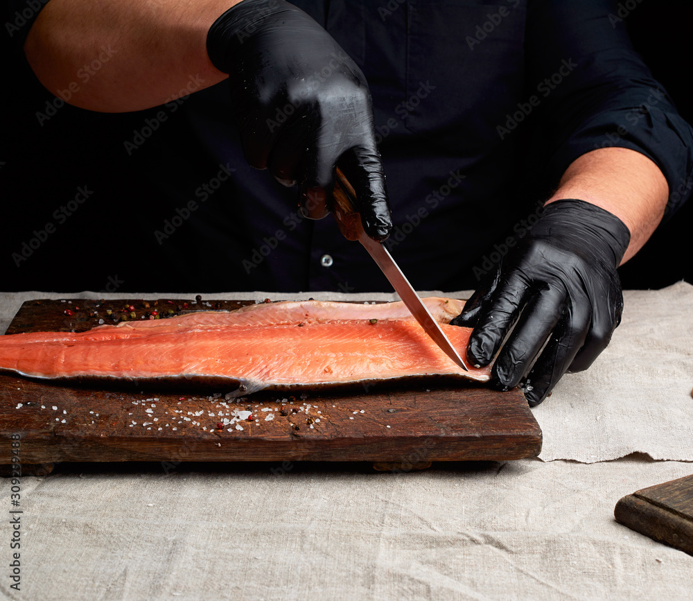 chef in black clothes and black latex gloves cuts fresh salmon filet into pieces
