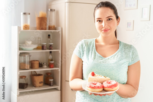 Body Care. Chubby girl standing in kitchen near fridge holding plate with dessert smiling friendly