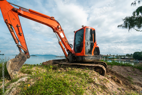 Small orange excavator on a ground against blue sky and sea for a works on construction site. Small tracked excavator standing on a ground with a blue sea on background. Heavy industry.