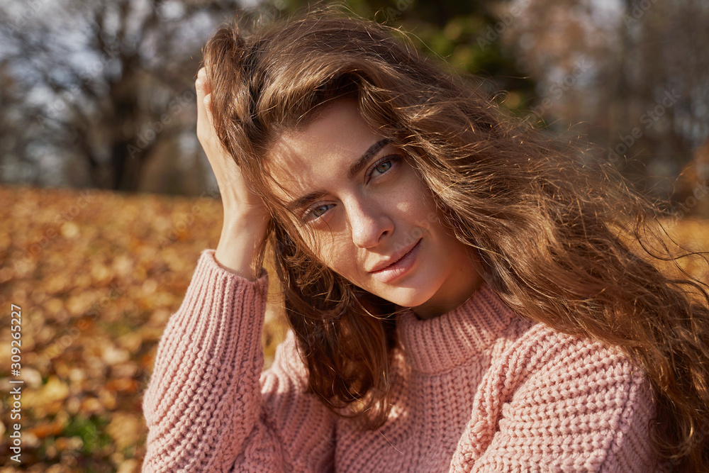 Sensual lady touches heads. Portrait of young woman with brown hair, green eyes wearing pink sweater sits in colorful forest or Park. Girl looking into camera has natural beauty and smiles tenderly. Sensual lady touches heads. Portrait of young woman with brown hair, green eyes wearing pink sweater sits in colorful forest or Park. Girl looking into camera has natural beauty and smiles tenderly.