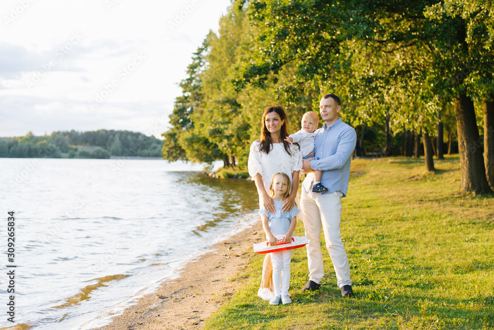 Fototapeta premium Friendly stylish beautiful family with two children standing on the shore of the lake. The girl is holding a boat. The concept of family holidays and vacations. Copy space