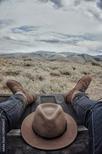Man with jeans and boots sitting on a truck bed with hat between legs and prarie in the background