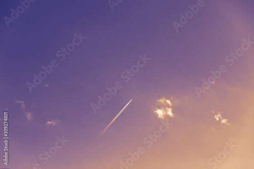 Small clouds and airplane white track or plane line on a blue sky background, copy space. Violet orange toned
