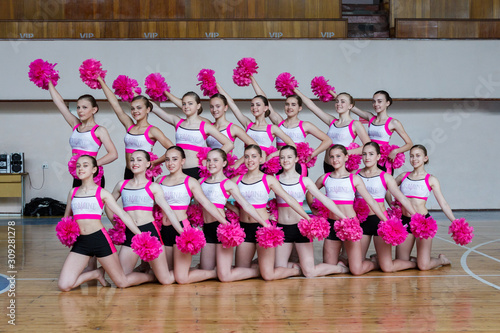 attractive young cheerleaders working out in sports club, group of cheerleaders with pom-poms in their hands, hands raised up, girls in black and pink suit with pompons on the background of the gym