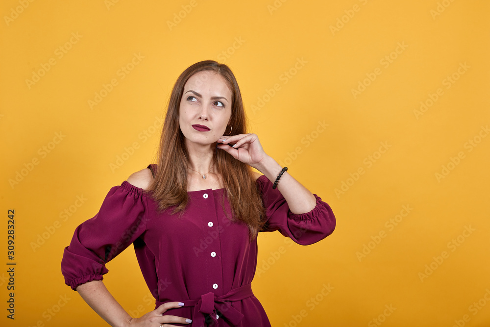 Young woman in burgundy bluse, over isolated orange background ponders idea holding hand on chin Young woman in burgundy bluse, over isolated orange background ponders idea holding hand on chin