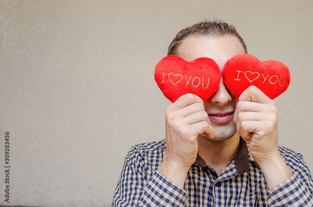 attractive dark-haired man with a smile, with two small red hearts in his hands, covering his eyes with paper heart symbols, standing on a light background. Valentine's day attractive dark-haired man with a smile, with two small red hearts in his hands, covering his eyes with paper heart symbols, standing on a light background. Valentine's day
