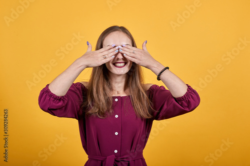 Ginger woman over isolated orange background in burgundy bluse with bow covering eyes by hands with smile on face. © Petro