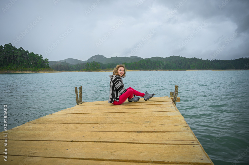 Young girl smiling sitting on a pier of a lagoon with mountains in the background and cloudy sky Young girl smiling sitting on a pier of a lagoon with mountains in the background and cloudy sky
