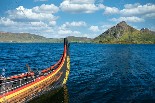 Viking warrior ancient  wooden long ship on sea with blue cloudy sky and green mountain in the background. Vikings, war equipment and history concept. © Jon Anders Wiken
