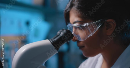 Female research scientist looking into microscope, wearing safety glasses.Blue lighting in a dark lab room.Close up, slider, shot with BMPCC 4K.Biochemistry, pharmaceutical medicine, science concept photo