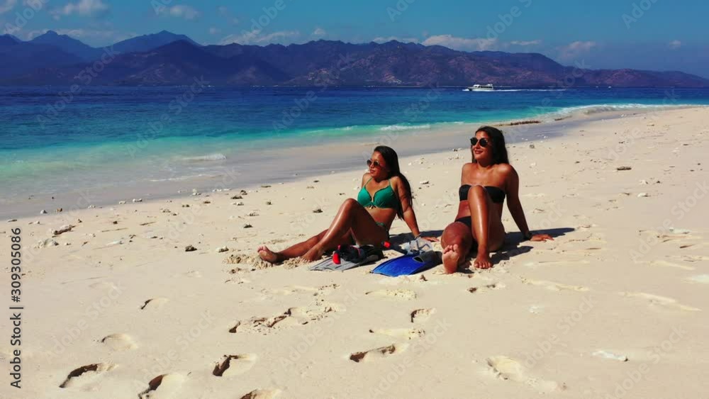 Beautiful girls tanning on white sandy beach under hot sun rays of tropic, alongside blue turquoise sea with white waves splashing on shore of tropical island in Bali Beautiful girls tanning on white sandy beach under hot sun rays of tropic, alongside blue turquoise sea with white waves splashing on shore of tropical island in Bali