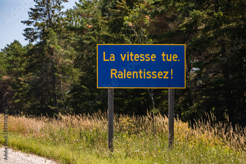Blue Information Road Sign with french yellow writing, Speed kills. Slow down !, on rural country roadside, forest trees background, Ontario, Canada