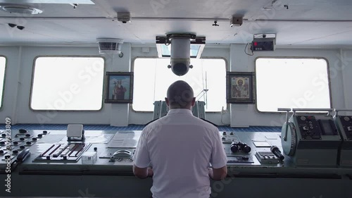 back view of captain of the cargo ship standing by the dashboard and steering wheel. russian man controls transport. concept of business, job, icon, cabin, deck.