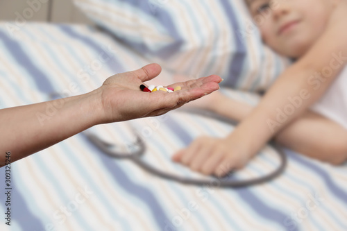 female hand holds out, offering, a handful of colored pills and capsules, close-up, blurred image of a lying child in the background, copy space, concept of medical care, treatment © kittyfly