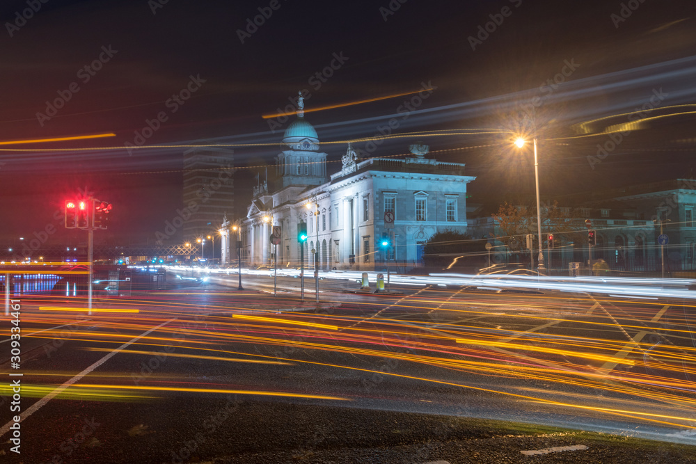 Fototapeta premium Niewyraźne ruchowe długie linie naświetlania światła reflektorów samochodowych w nocy. Widok ulicy na centrum Dublina z Urzędem Celnym w tle w Dublinie, Irlandia.