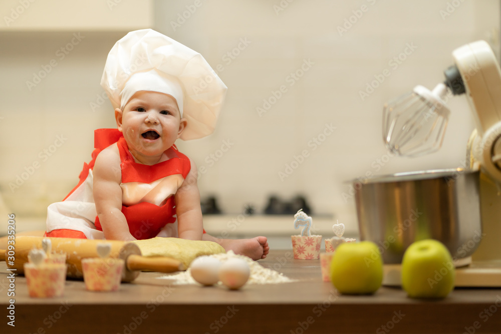 A Child, Baby, Girl, sits On The Kitchen Table, In A Cook's Cap A Child, Baby, Girl, sits On The Kitchen Table, In A Cook's Cap