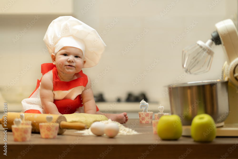 A Child, Baby, Girl, sits On The Kitchen Table, In A Cook's Cap A Child, Baby, Girl, sits On The Kitchen Table, In A Cook's Cap
