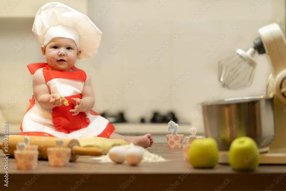 A Child, Baby, Girl, sits On The Kitchen Table, In A Cook's Cap A Child, Baby, Girl, sits On The Kitchen Table, In A Cook's Cap