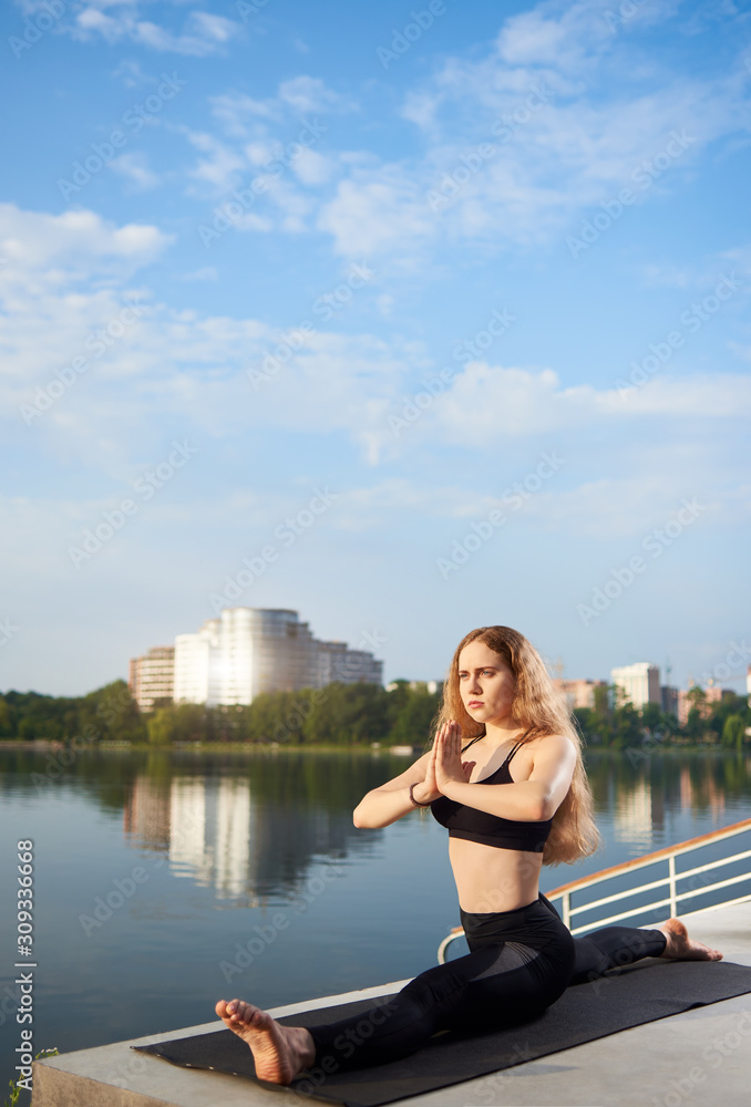 Young woman practicing yoga on urban lake, wearing black top and pants. Relax in nature under blue sky. Sitting on the twine. Meditation. Residential background. Copy space Young woman practicing yoga on urban lake, wearing black top and pants. Relax in nature under blue sky. Sitting on the twine. Meditation. Residential background. Copy space