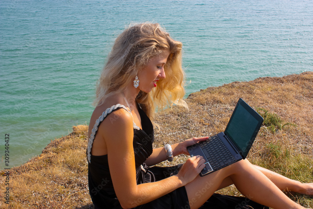 blonde with long hair and in a black dress sits on the edge of a cliff, on the seascape, with a laptop