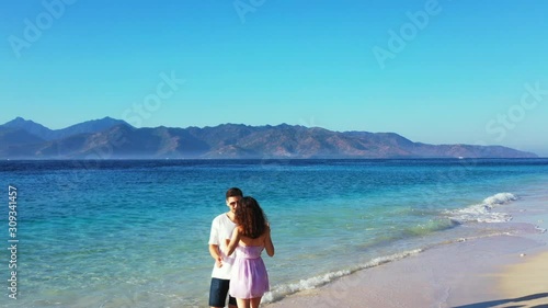 Couple hugging and kissing in front of blue sea with white waves washing exotic beach on a bright afternoon with mountains and clear sky background