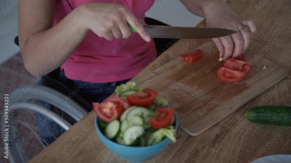 Disabled american woman cooking vegetarian salad sitting in wheelchair at table in home. Handicapped female cutting fresh tomato holding knife in hand at wooden desk in kitchen interior. Concept Disabled american woman cooking vegetarian salad sitting in wheelchair at table in home. Handicapped female cutting fresh tomato holding knife in hand at wooden desk in kitchen interior. Concept