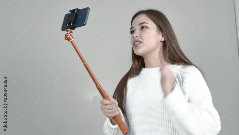 Grey background Caucasian girl white jumper Straight long hair, Holding orange selfie stick, looking at camera, laughing, talking animatedly, waving Grey background Caucasian girl white jumper Straight long hair, Holding orange selfie stick, looking at camera, laughing, talking animatedly, waving